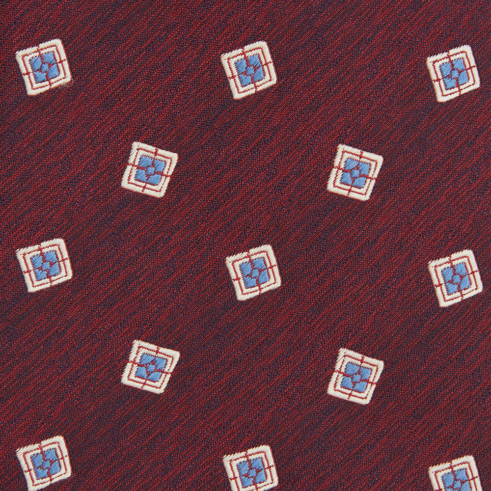 The Turnbull & Asser Burgundy Multi Geo Silk Tie features burgundy silk with blue, white, and yellow geometric designs—ideal for bold statement shirts. Shown rolled against a plain white background.