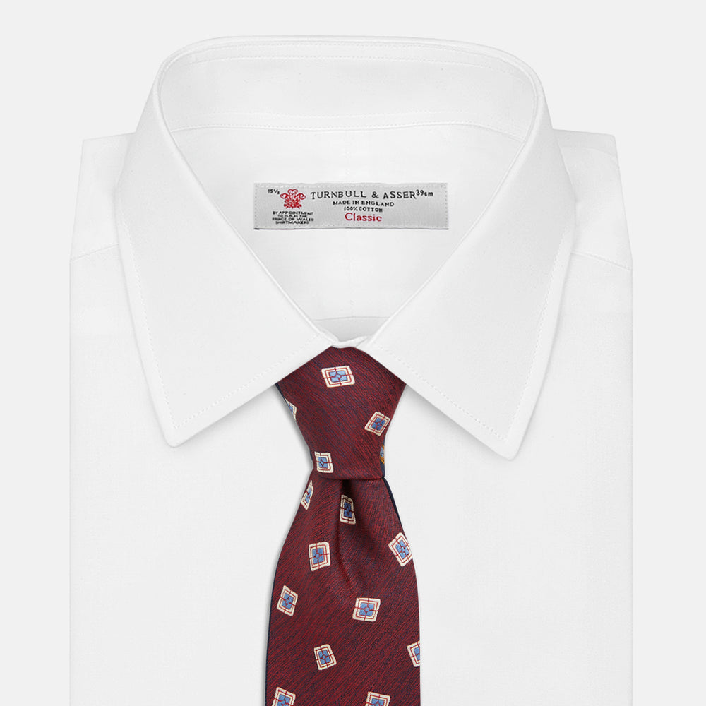 The Turnbull & Asser Burgundy Multi Geo Silk Tie features burgundy silk with blue, white, and yellow geometric designs—ideal for bold statement shirts. Shown rolled against a plain white background.