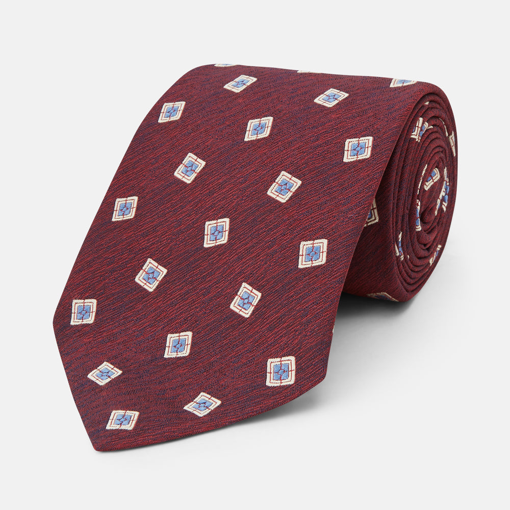 The Turnbull & Asser Burgundy Multi Geo Silk Tie features burgundy silk with blue, white, and yellow geometric designs—ideal for bold statement shirts. Shown rolled against a plain white background.