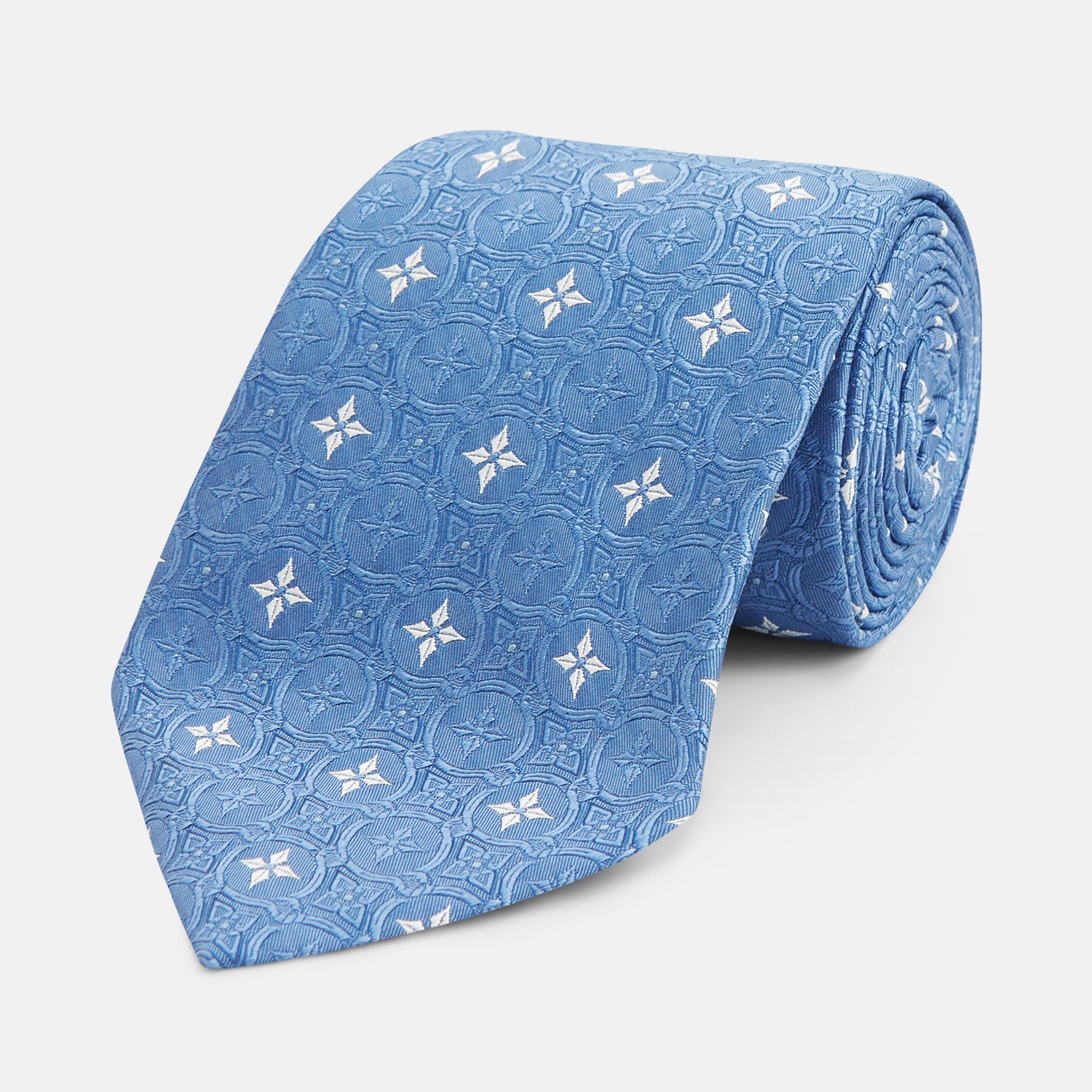 View 1 of product The Blue Multi Neat Silk Tie by Turnbull & Asser features a geometric pattern of white four-pointed shapes and circles, crafted in blue silk from the autumn-winter collection. Displayed rolled on a plain light background.