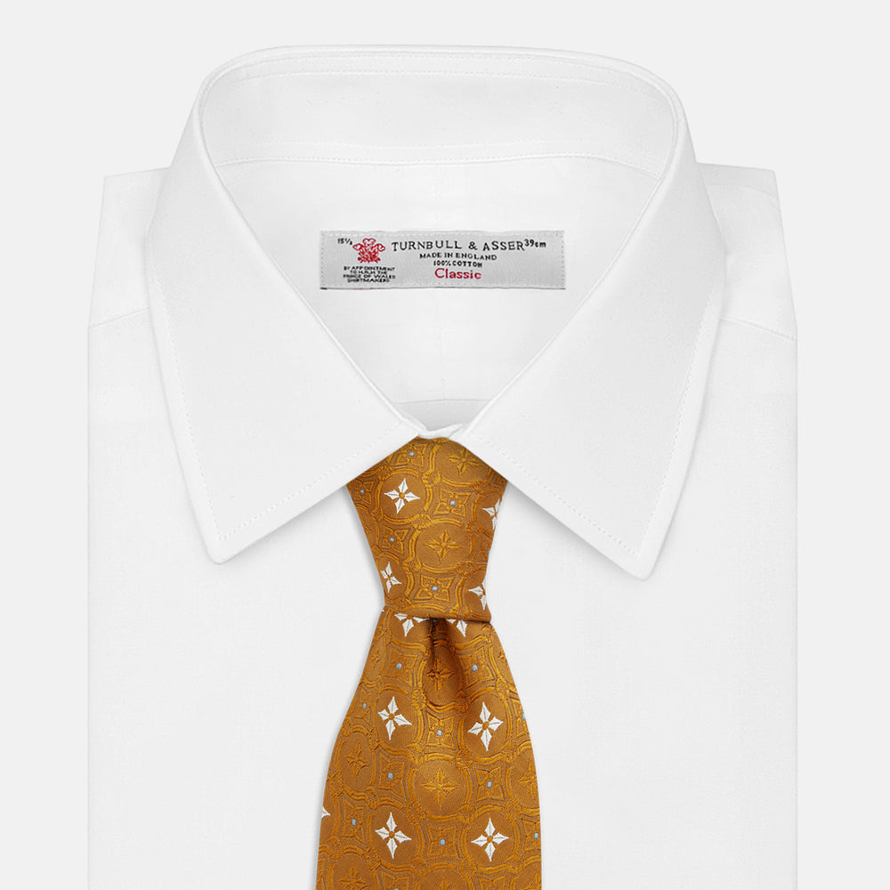 The Orange Multi Neat Silk Tie by Turnbull & Asser, from the autumn-winter collection, features a golden yellow geometric pattern on textured silk. Shown rolled on a white background, it pairs perfectly with bold signature shirts.