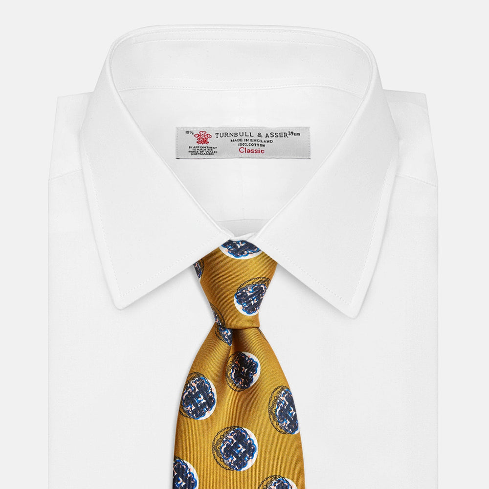 A rolled-up Turnbull & Asser Gold Shadow Spot Printed Silk Tie, featuring blue and white circular patterns on fine Adamley Macclesfield silk, displayed against a white background.