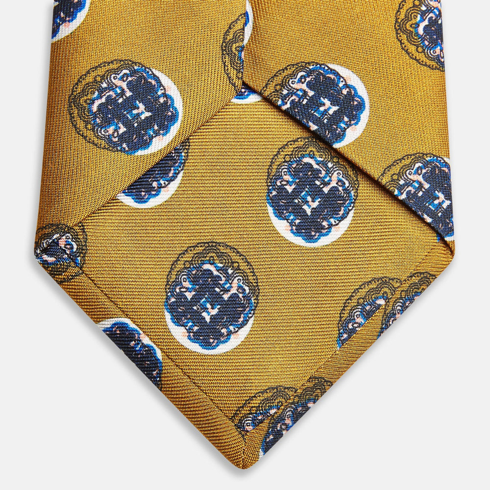 A rolled-up Turnbull & Asser Gold Shadow Spot Printed Silk Tie, featuring blue and white circular patterns on fine Adamley Macclesfield silk, displayed against a white background.
