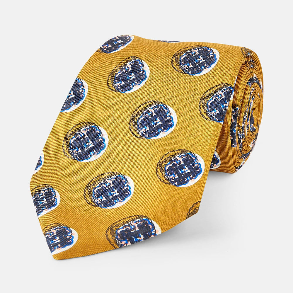 A rolled-up Turnbull & Asser Gold Shadow Spot Printed Silk Tie, featuring blue and white circular patterns on fine Adamley Macclesfield silk, displayed against a white background.
