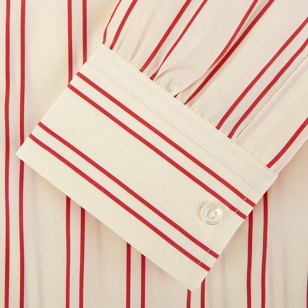 A man poses against a beige backdrop in the Turnbull & Asser Red and Off-White Stripe Cotton Weekend Fit Finch Shirt, styled with dark blue jeans and a brown belt for a timeless look. One hand rests in his pocket as he faces slightly to the side.