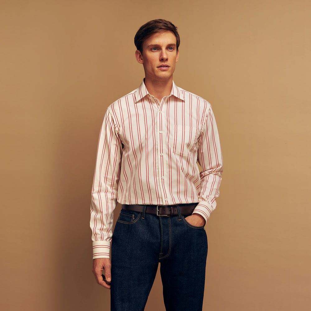 A man poses against a beige backdrop in the Turnbull & Asser Red and Off-White Stripe Cotton Weekend Fit Finch Shirt, styled with dark blue jeans and a brown belt for a timeless look. One hand rests in his pocket as he faces slightly to the side.
