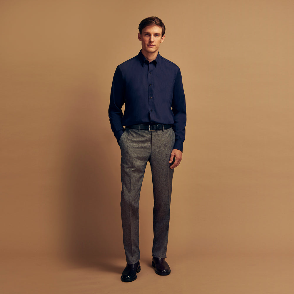 A man stands against a tan background, wearing the Turnbull & Asser Navy Wool Blend Weekend Fit Hayne Shirt tucked into gray dress pants with a black belt and shoes. He has short hair, a neutral expression, and his hands are in his pockets for a casual look.