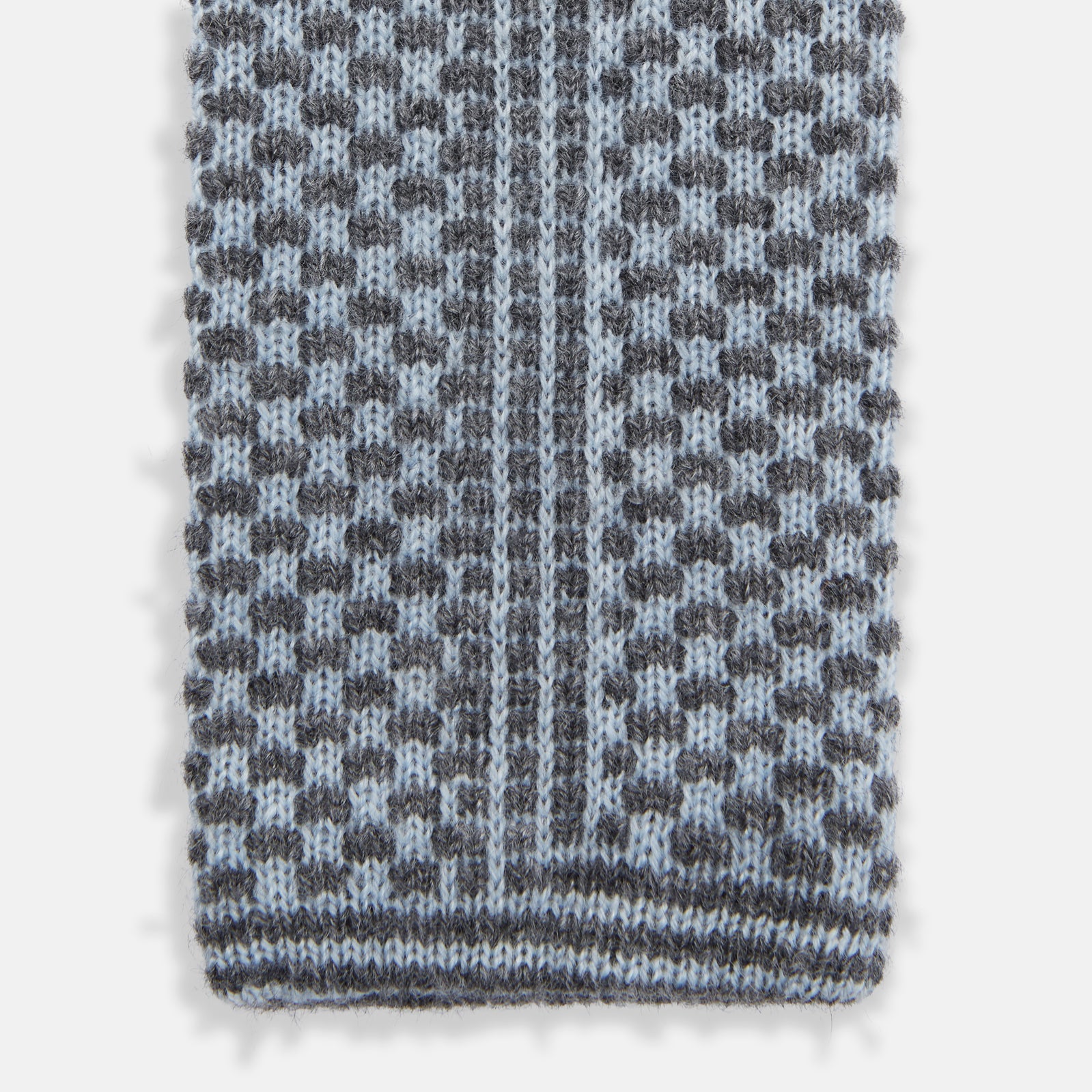 View 4 of product The Turnbull & Asser Pale Blue Multi Cashmere Knitted Tie features a subtle checkered pattern in black and light blue, making it a refined addition to your menswear collection. Shown rolled up on a plain white background.