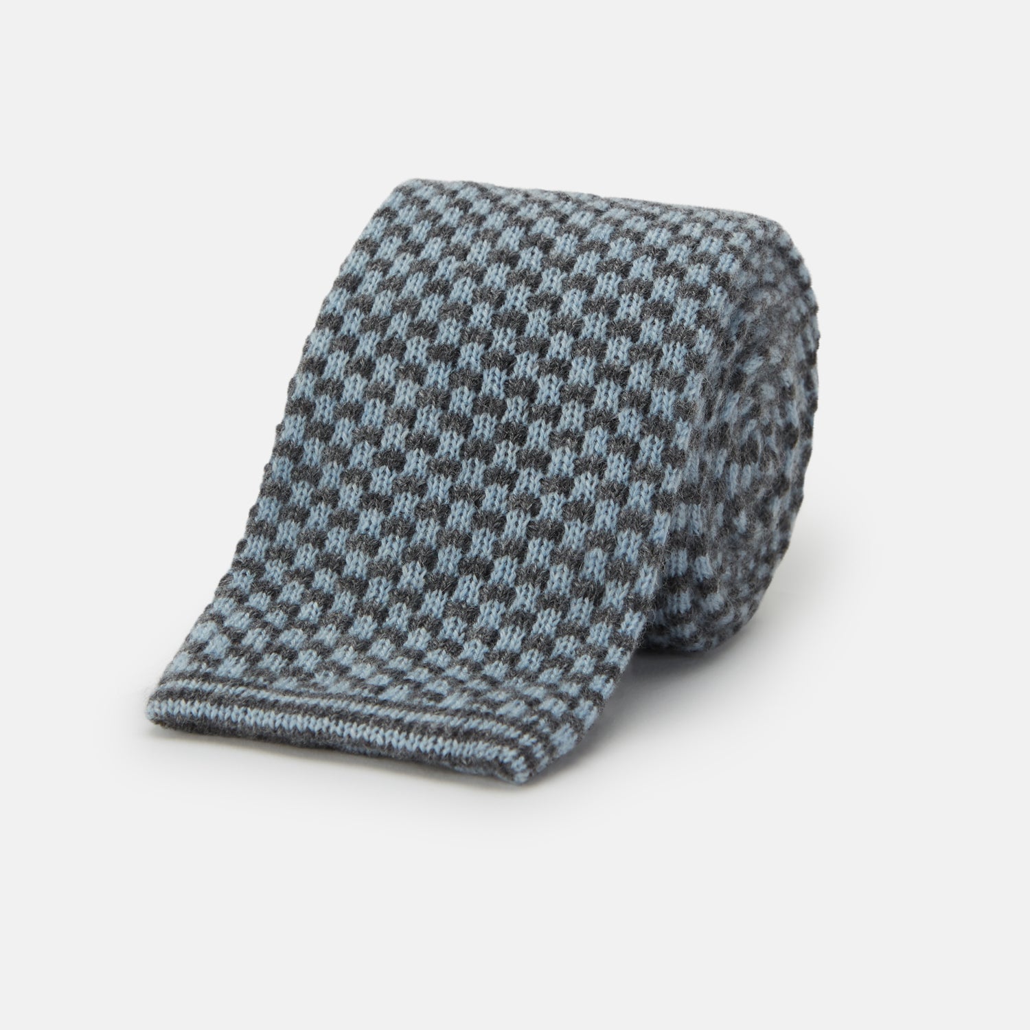 View 1 of product The Turnbull & Asser Pale Blue Multi Cashmere Knitted Tie features a subtle checkered pattern in black and light blue, making it a refined addition to your menswear collection. Shown rolled up on a plain white background.