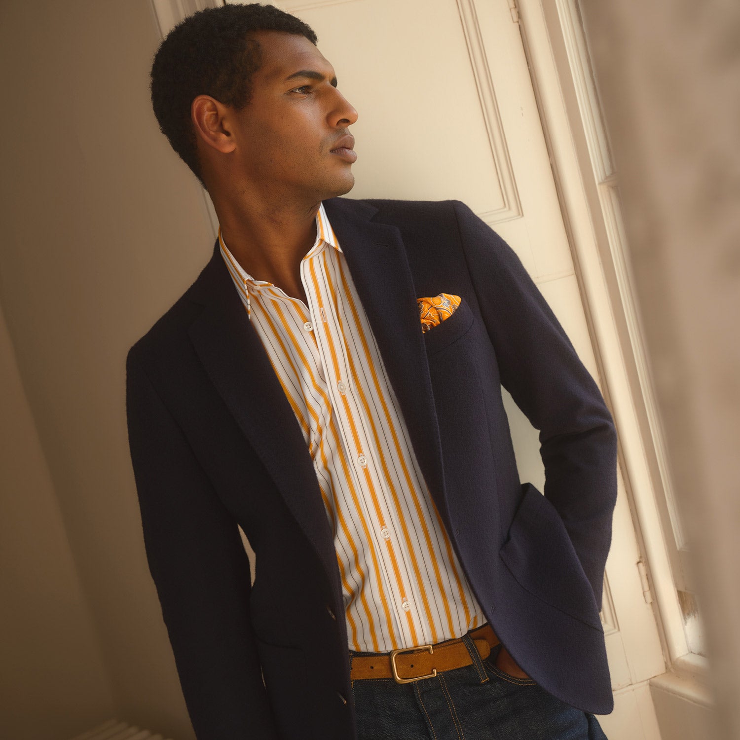 View 3 of product A man stands against a tan background wearing a Turnbull & Asser Orange Multi Stripe Regular Fit Shirt with T&A Collar and 3 Button Cuffs, paired with tan dress pants, brown belt, and brown shoes. He faces forward with a neutral expression.