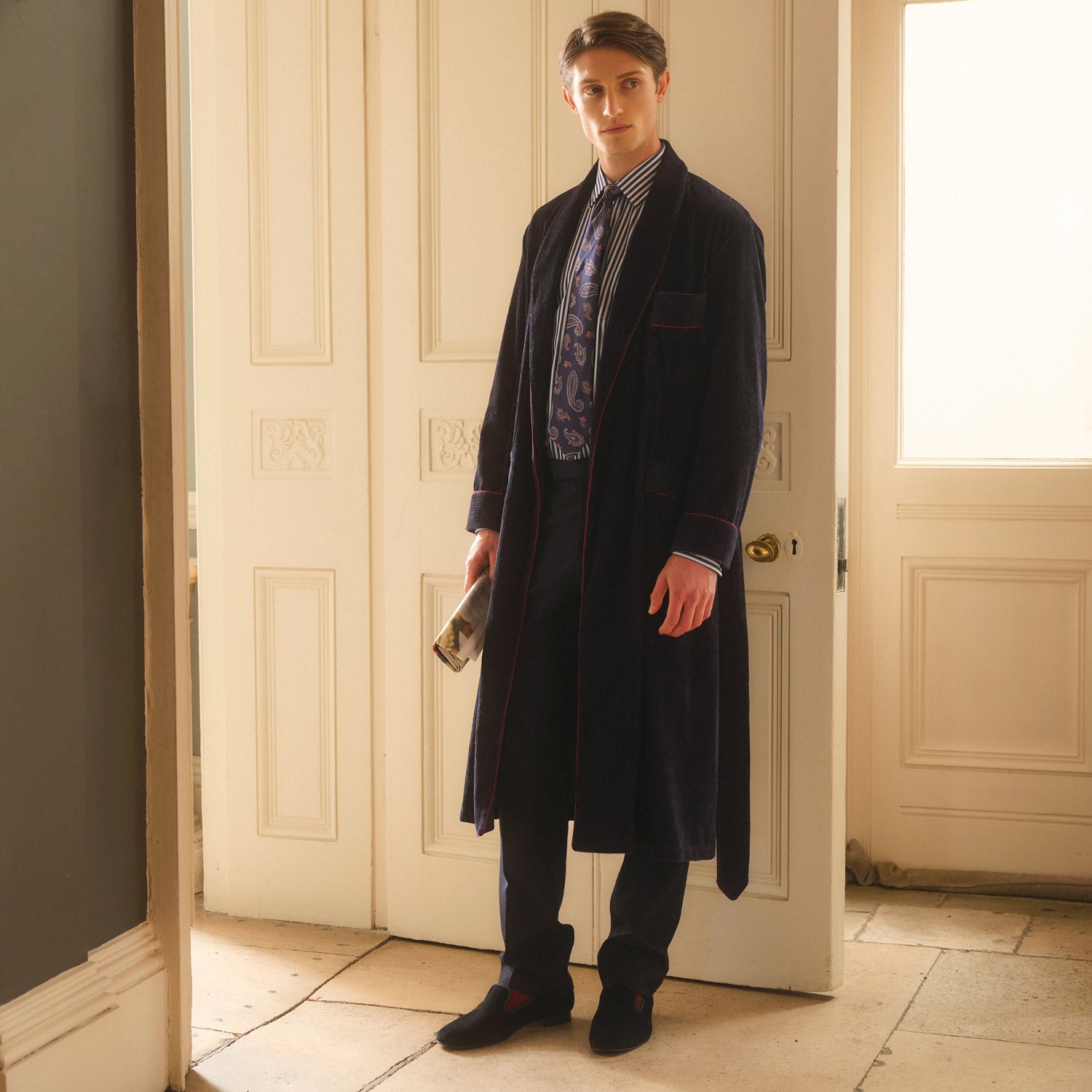 View 1 of product A man stands indoors before double doors, wearing the Turnbull & Asser Navy Linen & Cotton Cord Gown over a striped shirt and patterned tie, holding a book as soft natural light fills the scene.