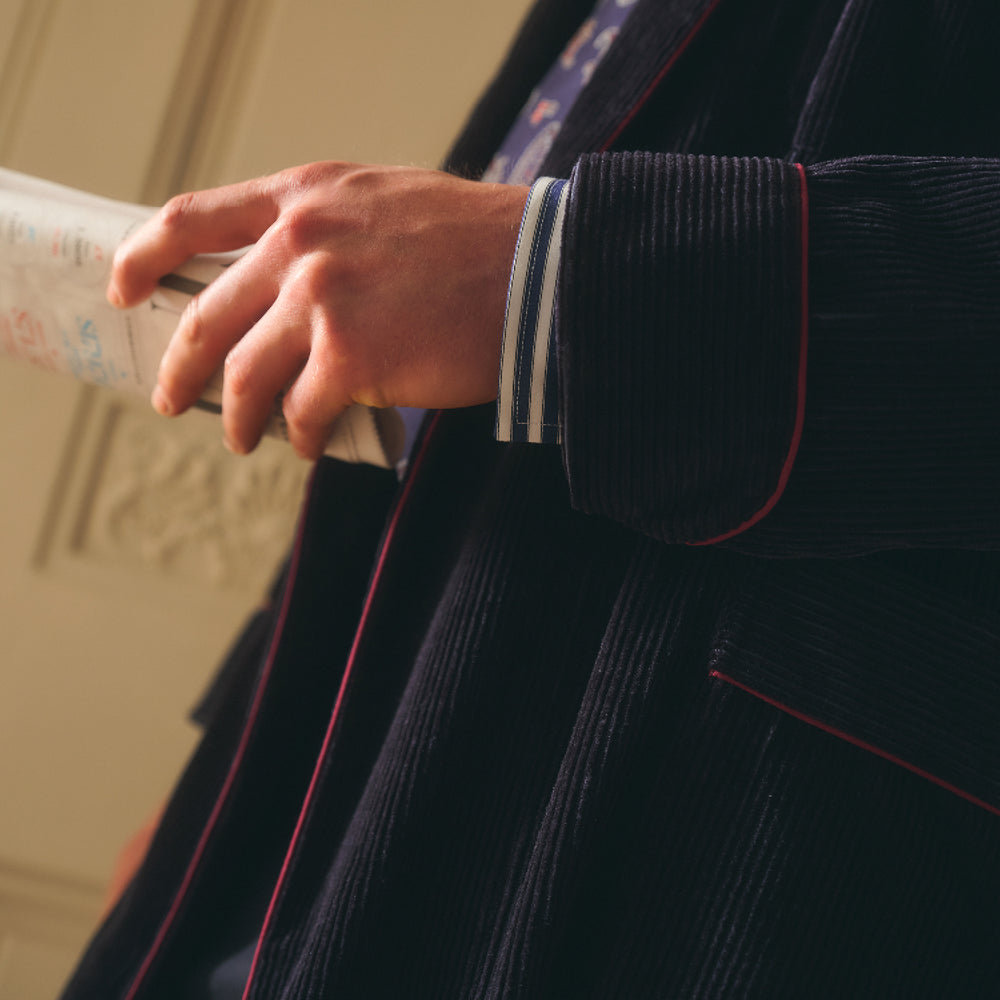 A man stands indoors before double doors, wearing the Turnbull & Asser Navy Linen & Cotton Cord Gown over a striped shirt and patterned tie, holding a book as soft natural light fills the scene.