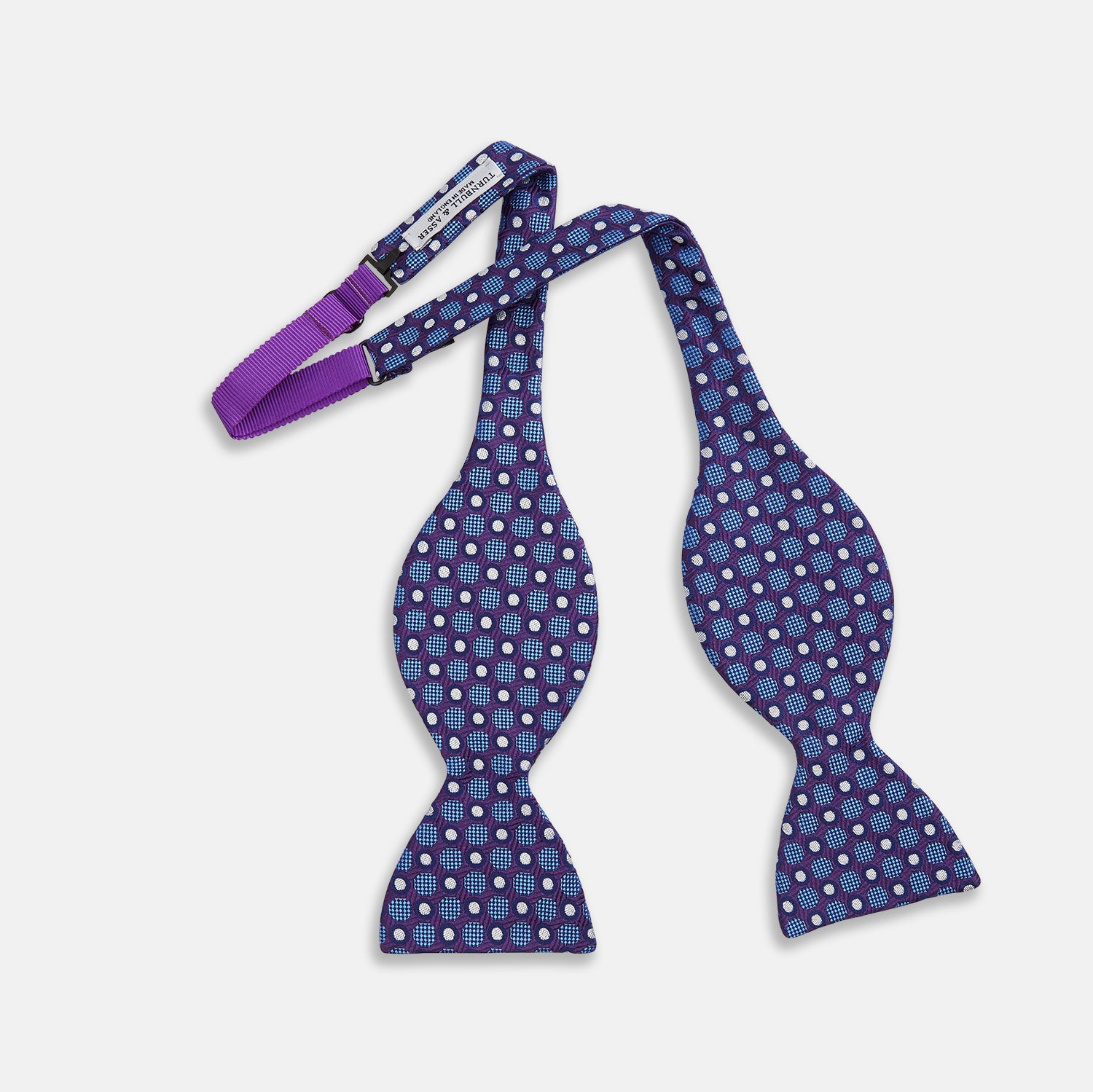 View 2 of product The Turnbull & Asser Plum and Blue Circle and Spot Silk Bow Tie, featuring blue with white and light blue dots on a purple background, is displayed on a plain white surface.