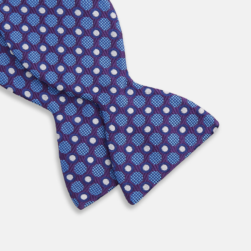 The Turnbull & Asser Plum and Blue Circle and Spot Silk Bow Tie, featuring blue with white and light blue dots on a purple background, is displayed on a plain white surface.