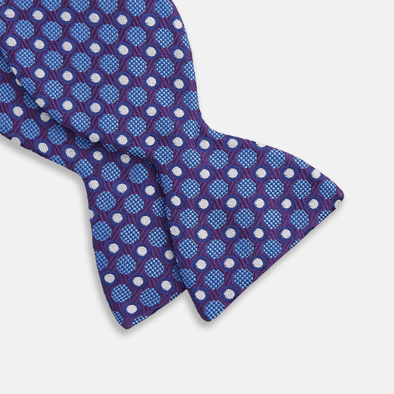View 3 of product The Turnbull & Asser Plum and Blue Circle and Spot Silk Bow Tie, featuring blue with white and light blue dots on a purple background, is displayed on a plain white surface.