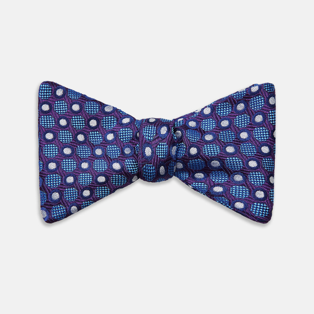 The Turnbull & Asser Plum and Blue Circle and Spot Silk Bow Tie, featuring blue with white and light blue dots on a purple background, is displayed on a plain white surface.