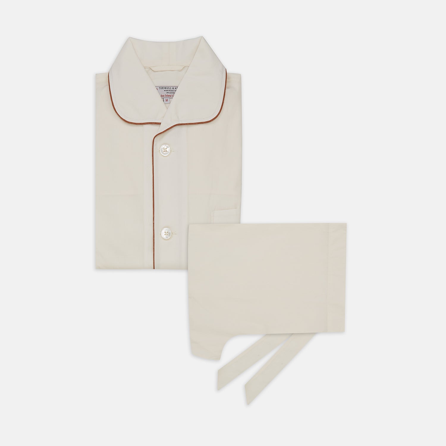 View 1 of product The Turnbull & Asser Cream Sea Island Cotton Pyjama Set features folded cream pyjamas with brown piping and a matching sleep mask with fabric ties, all elegantly displayed against a plain white background.