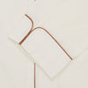 View 2 of product The Turnbull & Asser Cream Sea Island Cotton Pyjama Set features folded cream pyjamas with brown piping and a matching sleep mask with fabric ties, all elegantly displayed against a plain white background.