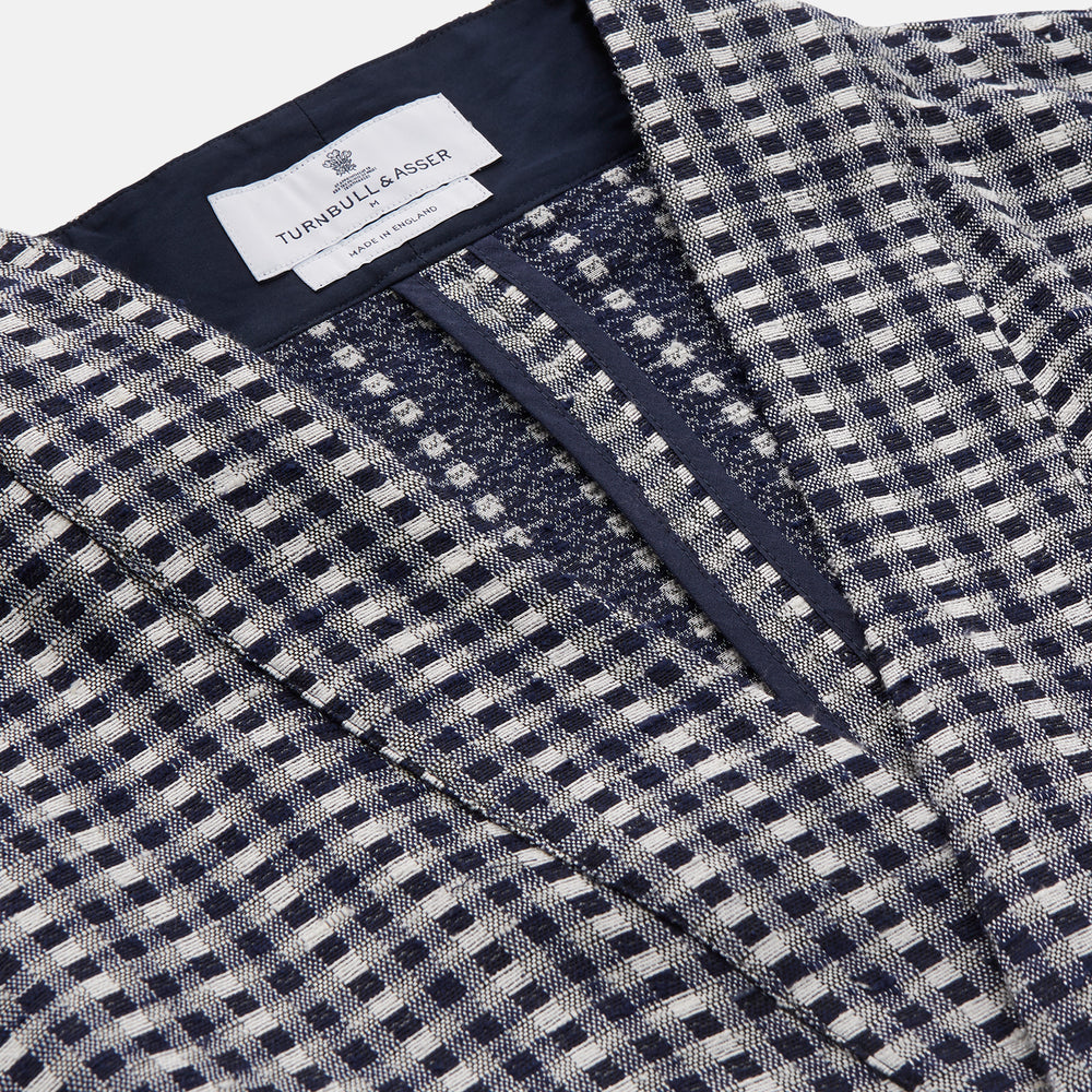 A barefoot person wears Turnbull & Asser's Navy Silk Chequered Kimono Gown with a belt over maroon pyjama sets, standing against a plain light background.