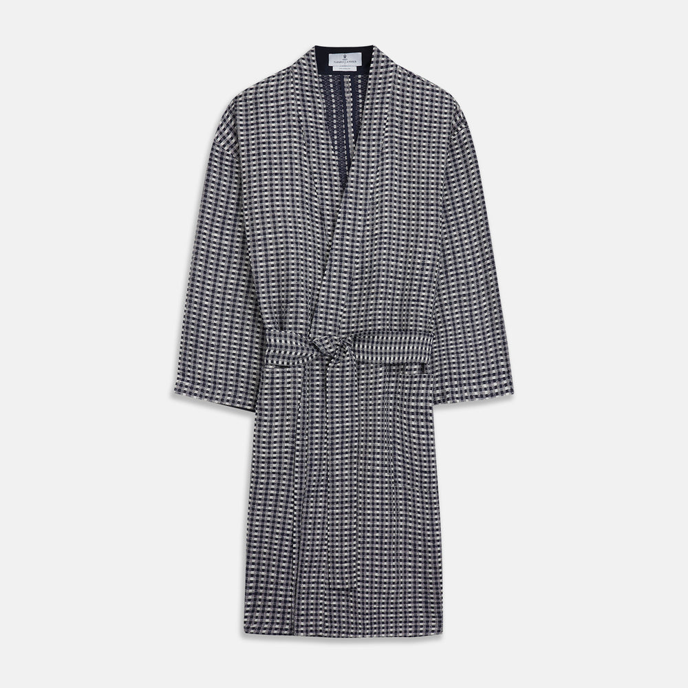 A barefoot person wears Turnbull & Asser's Navy Silk Chequered Kimono Gown with a belt over maroon pyjama sets, standing against a plain light background.