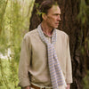 View 1 of product A man wearing a Turnbull & Asser Cream Abstract Stripe Print Lightweight Wool Scarf and a beige shirt stands outside by a tree, surrounded by greenery, gazing thoughtfully to the side.