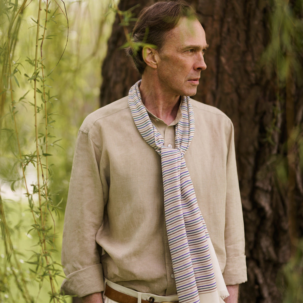 A man wearing a Turnbull & Asser Cream Abstract Stripe Print Lightweight Wool Scarf and a beige shirt stands outside by a tree, surrounded by greenery, gazing thoughtfully to the side.