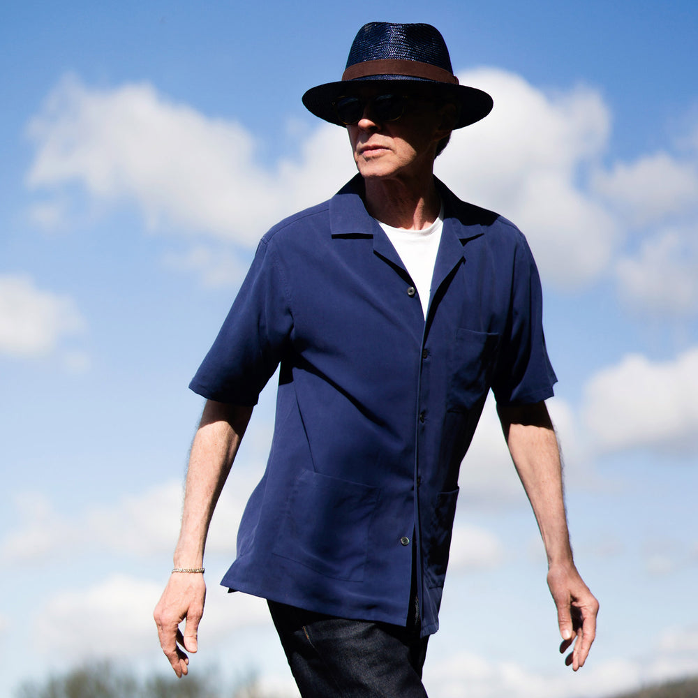 A man stands against a beige backdrop, wearing the Turnbull & Asser Navy Holiday Fit Silk Phillips Shirt with Revere Collar and dark pinstripe pants. He gazes downwards with a relaxed expression, embodying effortless summer style.