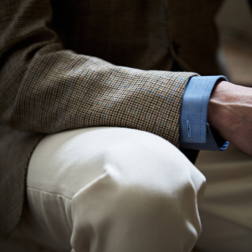 The Byron Wool Check Travel Jacket by Turnbull & Asser features a brown plaid pattern, notched lapel, four flap utility pockets, and dark buttons. This lightweight wool jacket is shown on a plain white background.