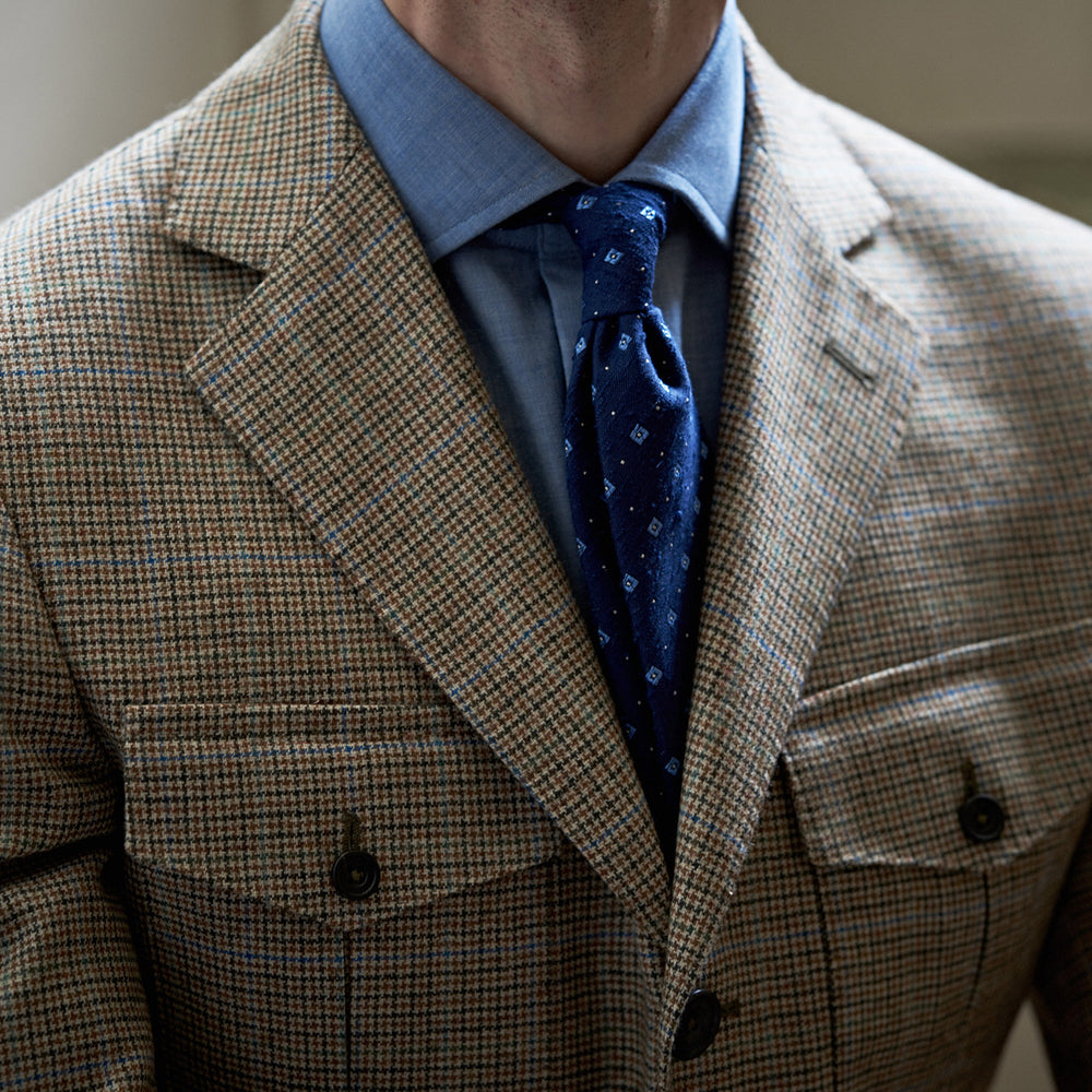 The Byron Wool Check Travel Jacket by Turnbull & Asser features a brown plaid pattern, notched lapel, four flap utility pockets, and dark buttons. This lightweight wool jacket is shown on a plain white background.