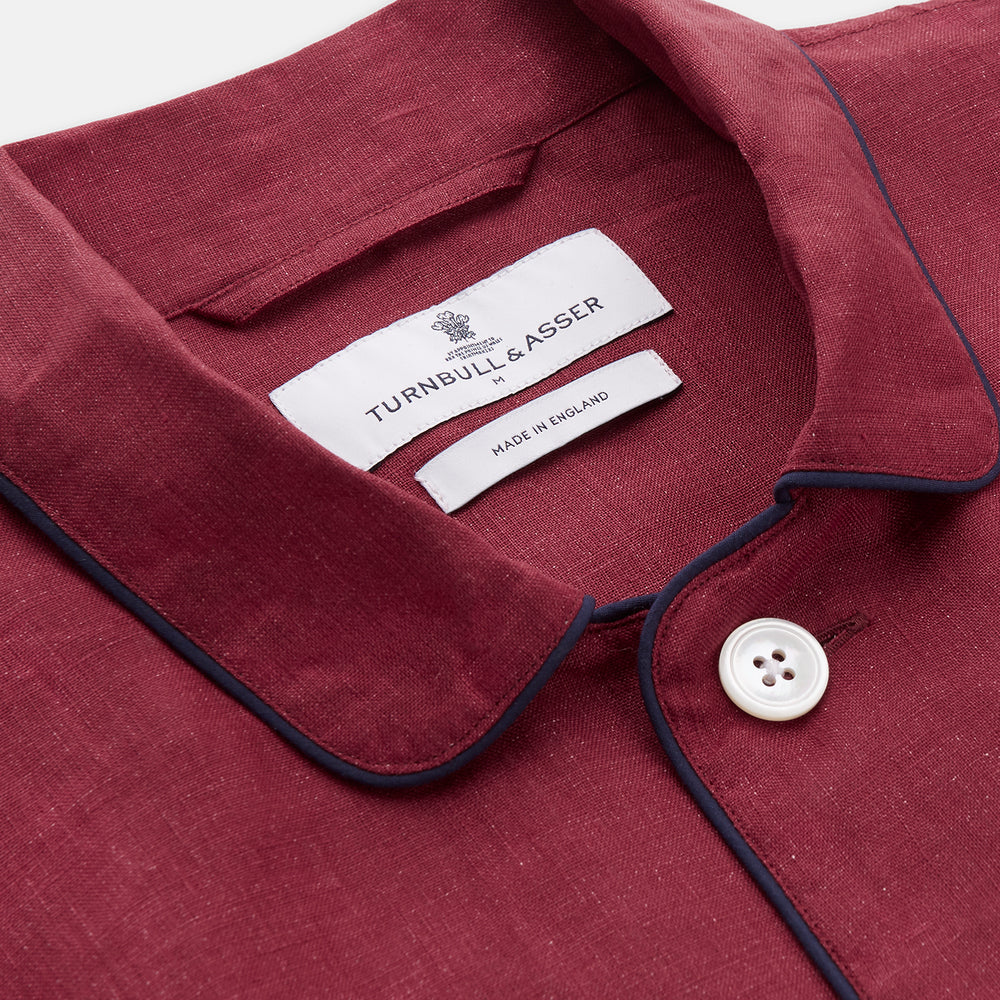 The Turnbull & Asser Burgundy Linen Modern Pyjama Set features a button-up, collared long-sleeve top with black piping and three pockets, plus matching pants with an elastic waistband.