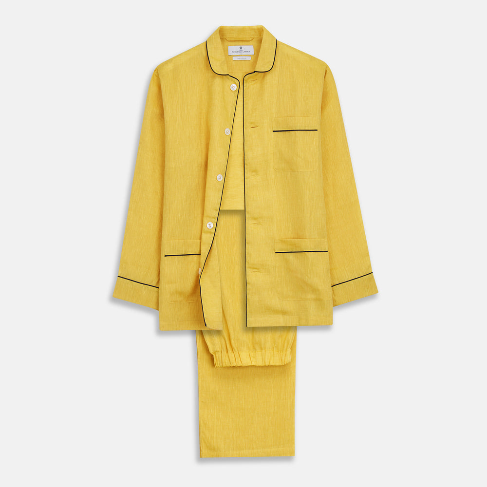 A barefoot person stands against a plain background, wearing the Turnbull & Asser Ochre Linen Modern Pyjama Set with black piping on the collar, cuffs, and pockets. The button-down top completes the look as they gaze to the side.