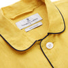 View 5 of product A barefoot person stands against a plain background, wearing the Turnbull & Asser Ochre Linen Modern Pyjama Set with black piping on the collar, cuffs, and pockets. The button-down top completes the look as they gaze to the side.