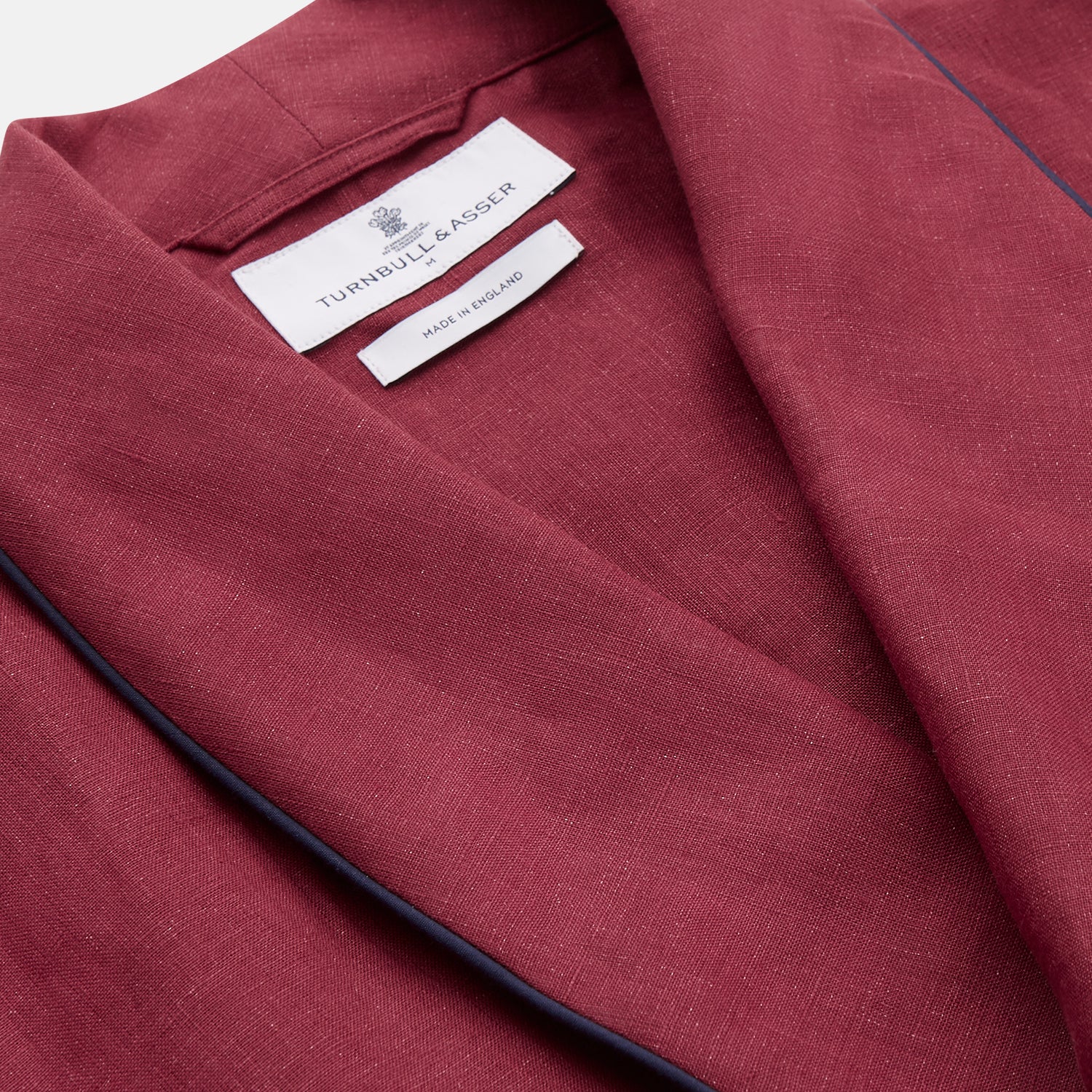 View 4 of product A person stands barefoot against a plain background, wearing the Turnbull & Asser Burgundy Linen Piped Gown belted at the waist over blue pajamas, looking to the side with one hand in the gown’s pocket.