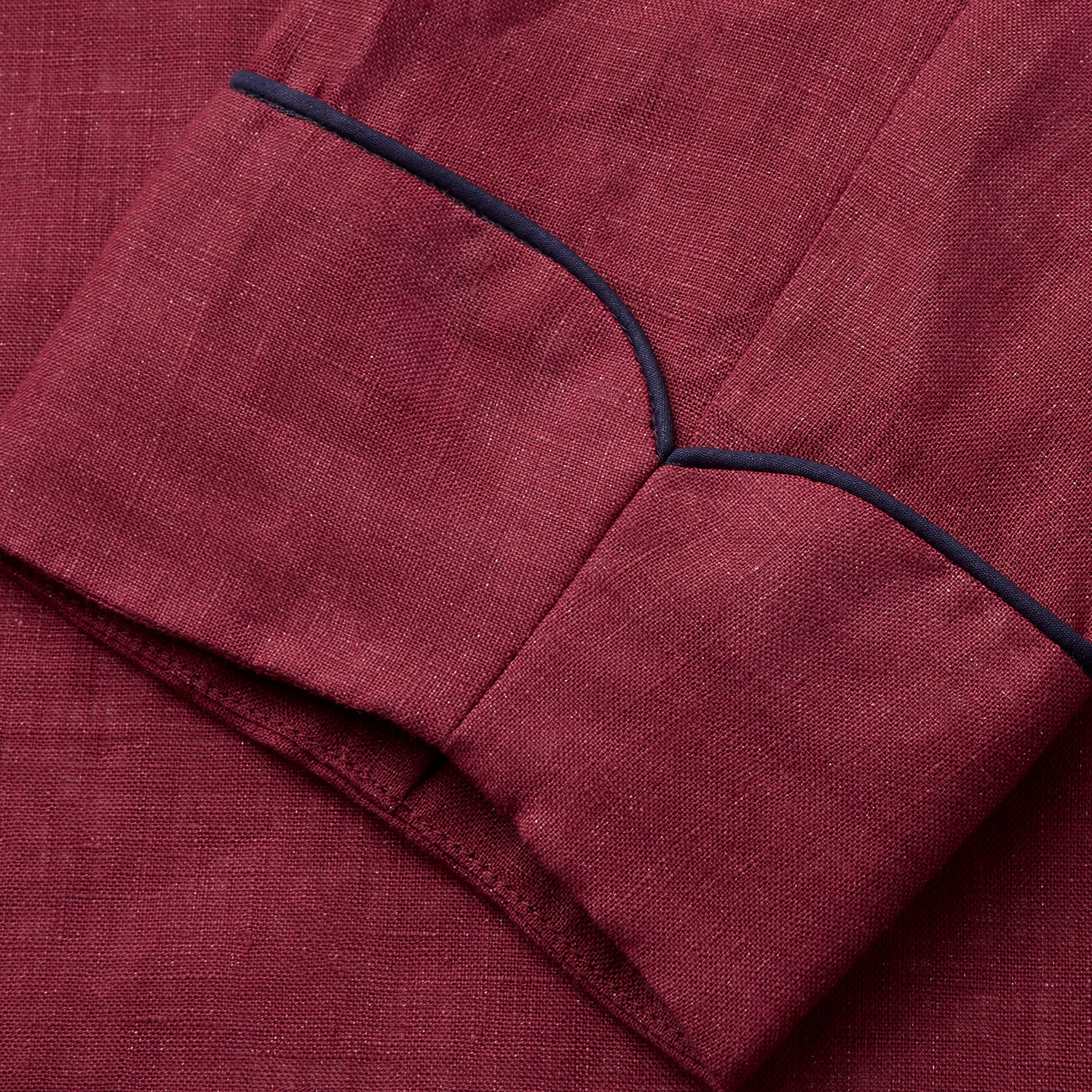 View 5 of product A person stands barefoot against a plain background, wearing the Turnbull & Asser Burgundy Linen Piped Gown belted at the waist over blue pajamas, looking to the side with one hand in the gown’s pocket.