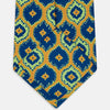 View 4 of product The Yellow Medallion Printed Silk Tie by Turnbull & Asser features a bold abstract medallion pattern in blue, green, and orange on yellow silk, displayed rolled up against a plain white background.
