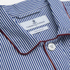 View 3 of product The Turnbull & Asser Navy & White Stripe Cotton Modern Pyjama Set features blue and white Bengal stripes with red piping, a long-sleeve button-up top, chest pocket, and matching pants. Shown laid flat on a white background.