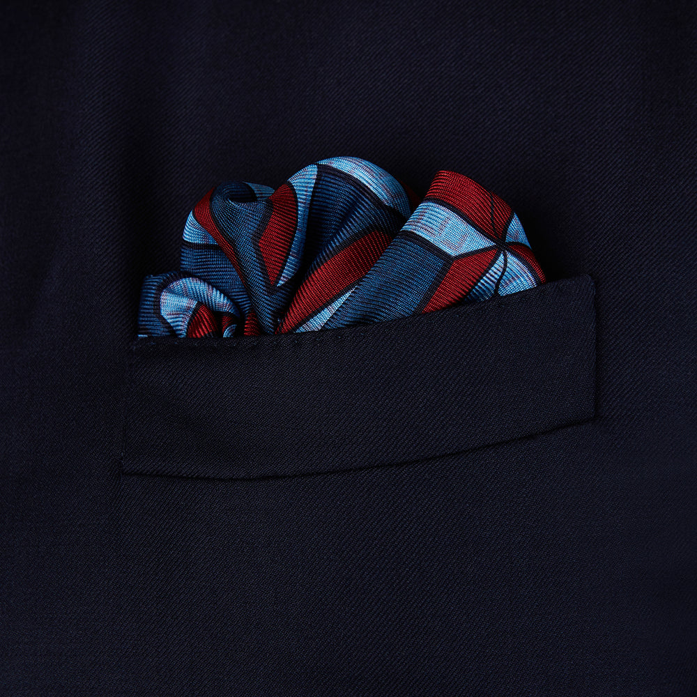 The Turnbull & Asser Red Multi Crosses Double-Faced Silk Pocket Square features red, white, and light blue Xs on dark blue silk with an orange border and the brand's name printed in the bottom right corner.