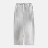 View 7 of product A person stands barefoot on a concrete floor, wearing the Turnbull & Asser Black & White Stripe Harriet Women’s Pyjama Set, featuring loose-fitting white pajamas with black vertical stripes, against a plain beige wall in soft side lighting.