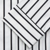 View 3 of product A person stands barefoot on a concrete floor, wearing the Turnbull & Asser Black & White Stripe Harriet Women’s Pyjama Set, featuring loose-fitting white pajamas with black vertical stripes, against a plain beige wall in soft side lighting.