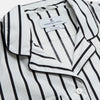 View 4 of product A person stands barefoot on a concrete floor, wearing the Turnbull & Asser Black & White Stripe Harriet Women’s Pyjama Set, featuring loose-fitting white pajamas with black vertical stripes, against a plain beige wall in soft side lighting.