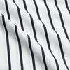 View 5 of product A person stands barefoot on a concrete floor, wearing the Turnbull & Asser Black & White Stripe Harriet Women’s Pyjama Set, featuring loose-fitting white pajamas with black vertical stripes, against a plain beige wall in soft side lighting.
