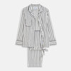 View 2 of product A person stands barefoot on a concrete floor, wearing the Turnbull & Asser Black & White Stripe Harriet Women’s Pyjama Set, featuring loose-fitting white pajamas with black vertical stripes, against a plain beige wall in soft side lighting.