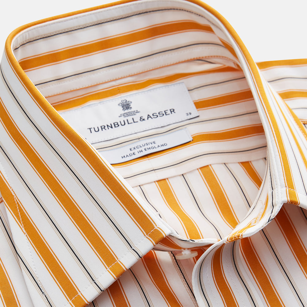 A man stands against a tan background wearing a Turnbull & Asser Orange Multi Stripe Regular Fit Shirt with T&A Collar and 3 Button Cuffs, paired with tan dress pants, brown belt, and brown shoes. He faces forward with a neutral expression.