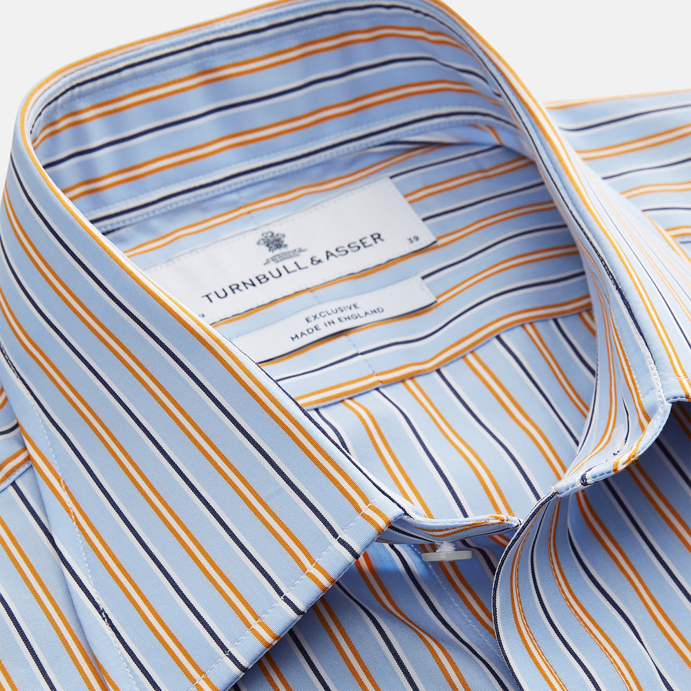 A man stands against a beige background, wearing a Turnbull & Asser Blue, Orange & Navy Multi Stripe Regular Fit Shirt with T&A collar and 3-button cuffs, tucked into tan dress pants with a black belt. He faces forward with a neutral expression.