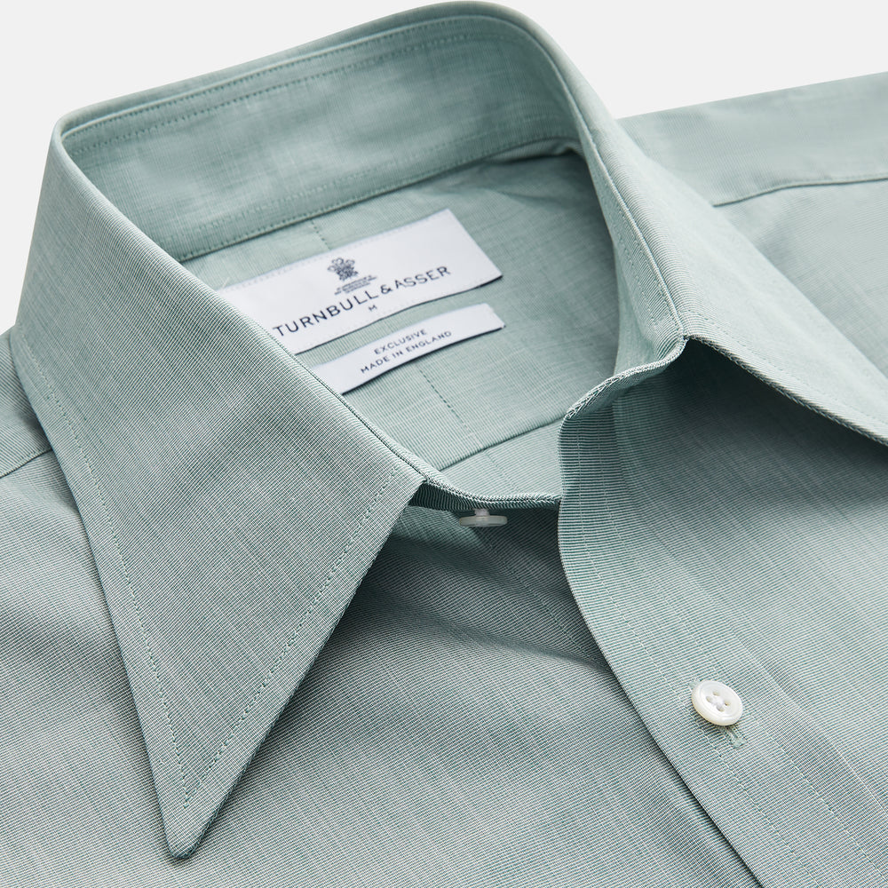 A man stands against a beige background wearing a Turnbull & Asser Green Weekend Fit Shirt with Long Point Collar and Single Button Cuffs, paired with a striped tie, dark trousers, and black dress shoes, hands in pockets and a neutral expression.