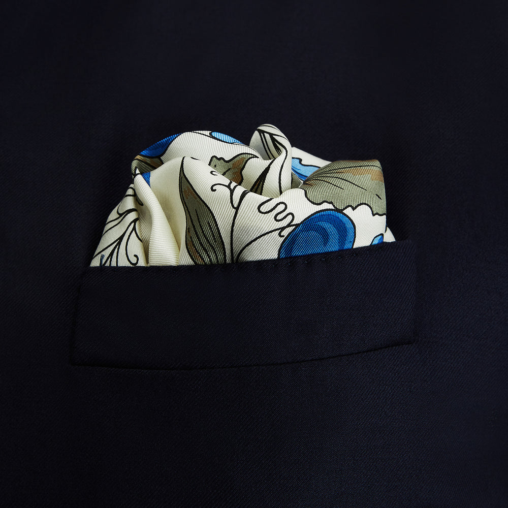 The Turnbull & Asser Pale Blue and Oat Foliage Pattern Silk Pocket Square is crafted in England, featuring a cream background with a circular blue and green floral design, bordered edges, and the brand's logo in one corner.