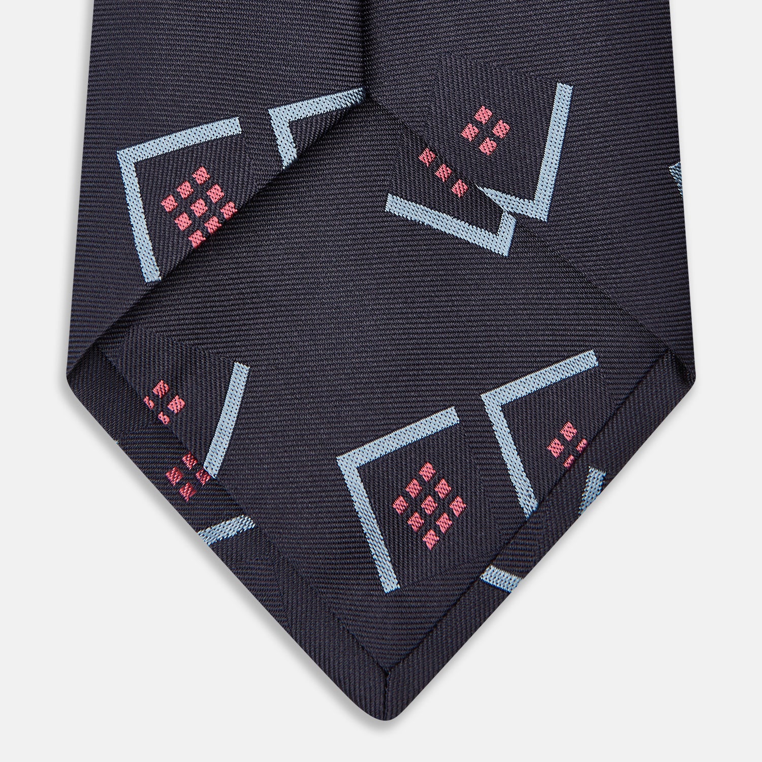 View 3 of product A Turnbull & Asser Navy Graphic Deco Silk Tie, featuring light blue chevrons and red squares on a dark navy background, shown rolled up against a plain white backdrop.