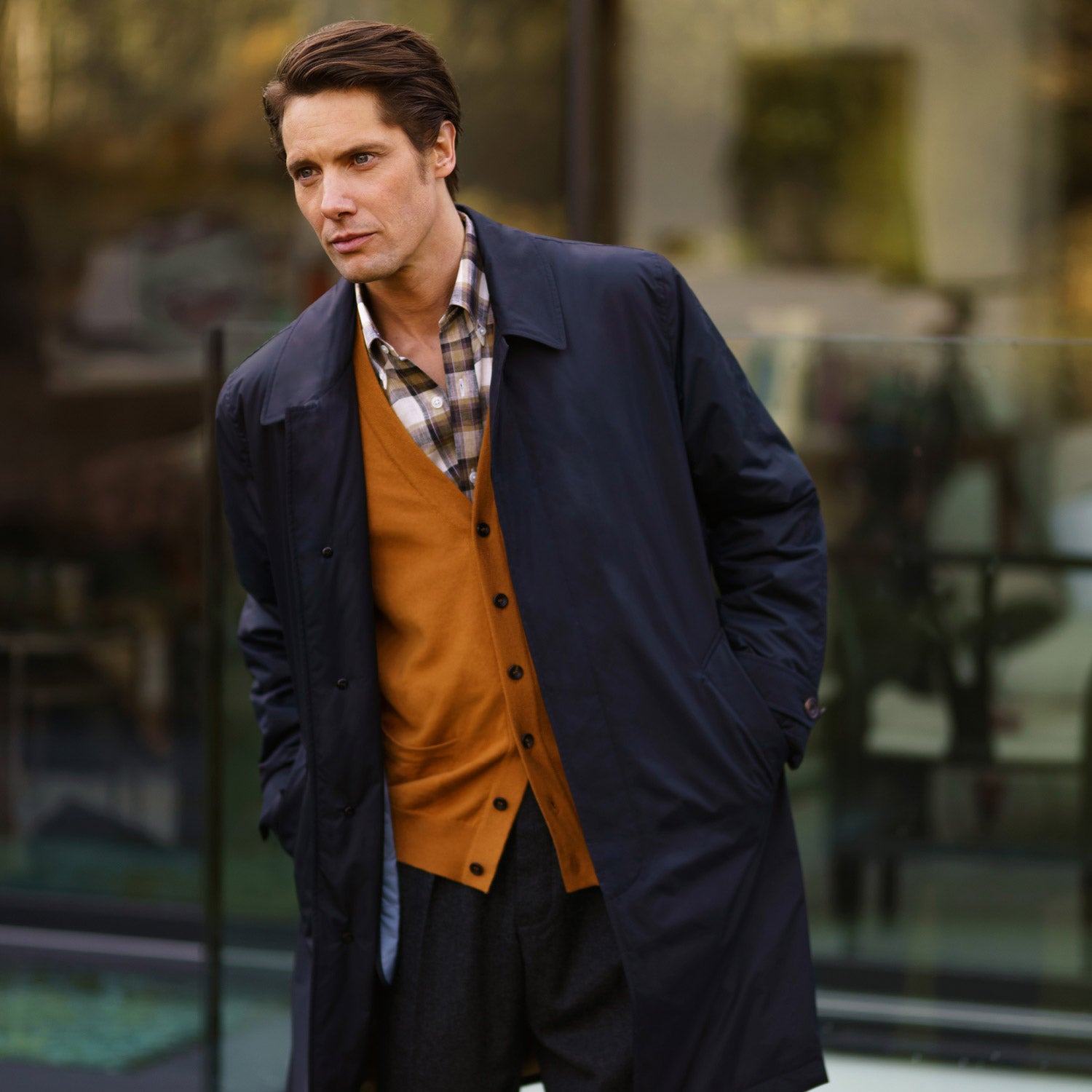 View 1 of product A man with brown hair stands outdoors, hands in his pockets, wearing a Turnbull & Asser Navy Sealup Quilted Liner Quintern Car Coat over a mustard cardigan. A glass barrier and blurred background are behind him.
