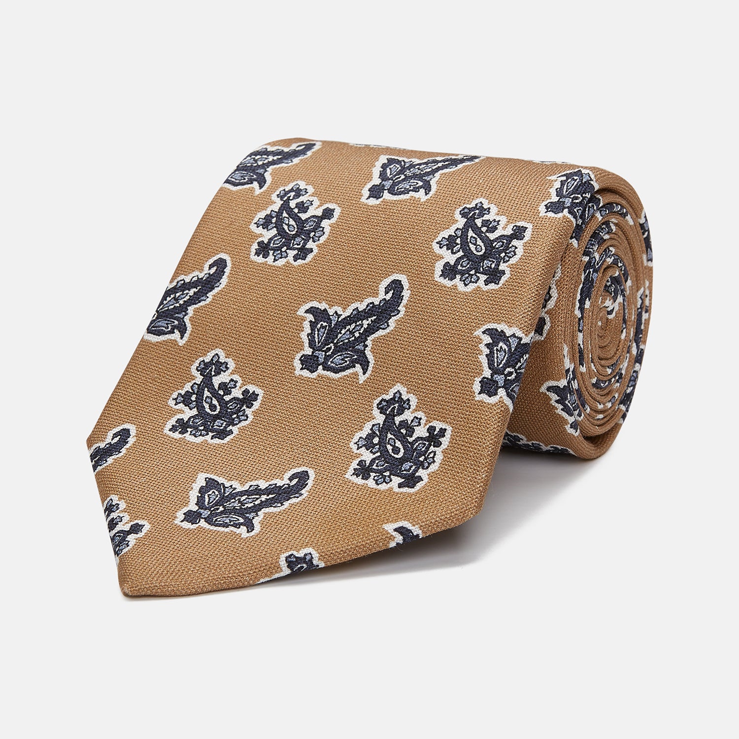 View 1 of product The Turnbull & Asser Toffee Paisley Floral Cotton Silk Blend Tie features a rolled-up design with dark blue and white paisley motifs on a tan background.