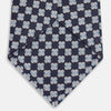 View 3 of product The Turnbull & Asser Indigo Graphic Floral Silk Jacquard Tie features a repeating pattern of light blue and white geometric shapes, shown rolled up on a plain white background.
